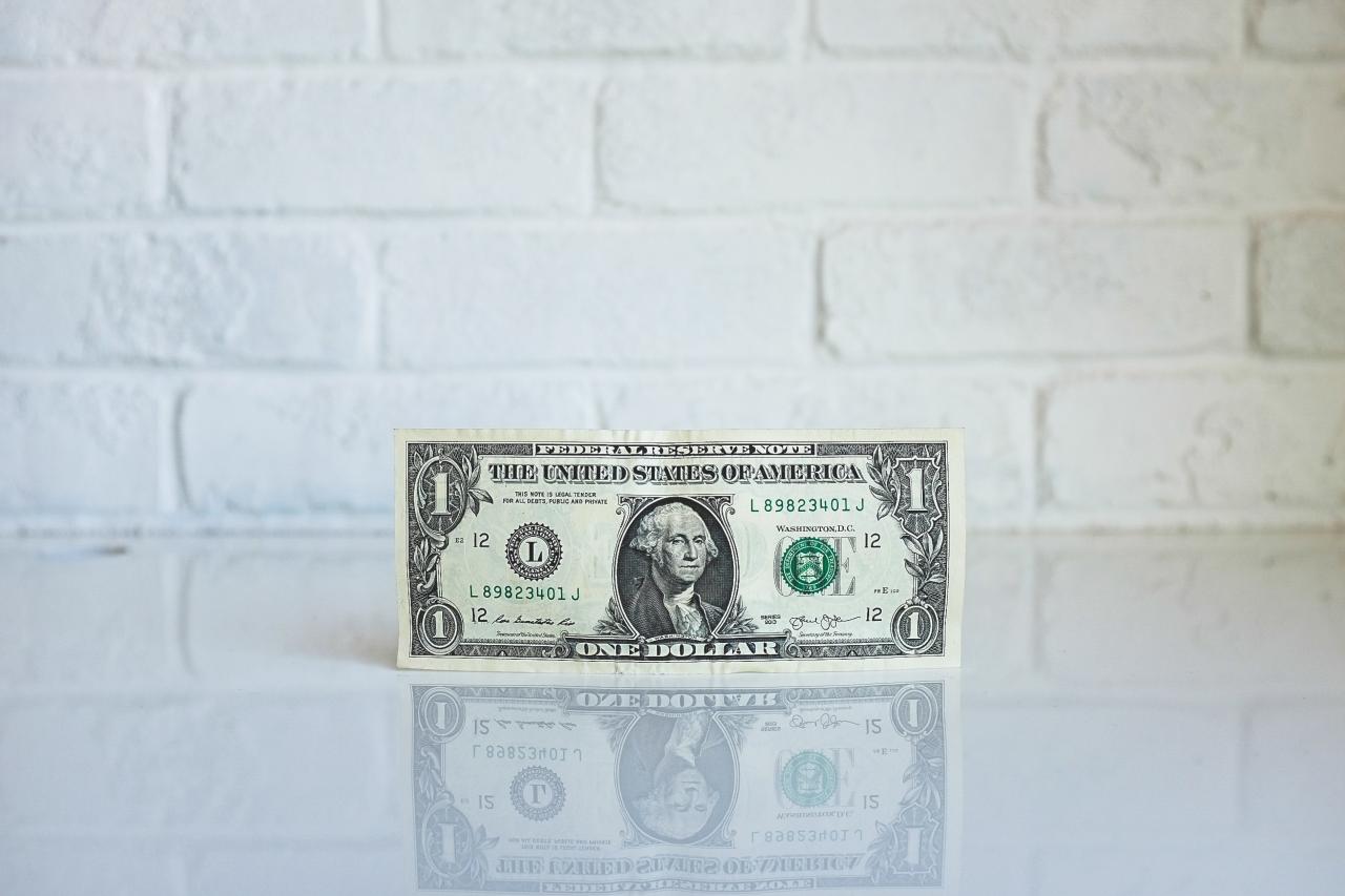 An image of a one dollar bill standing up against a white brick background