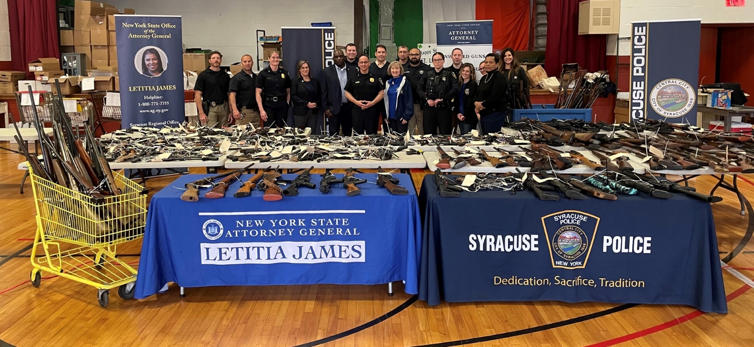 hundreds of guns on several table with a group of people behind them