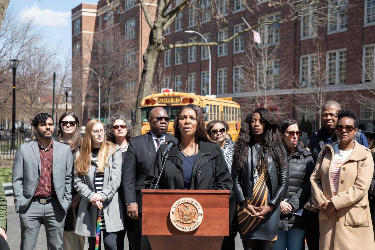 Attorney General Tish James stands in front of a podium. A crowd stands behind her, and in the background you can see a yellow school bus. 