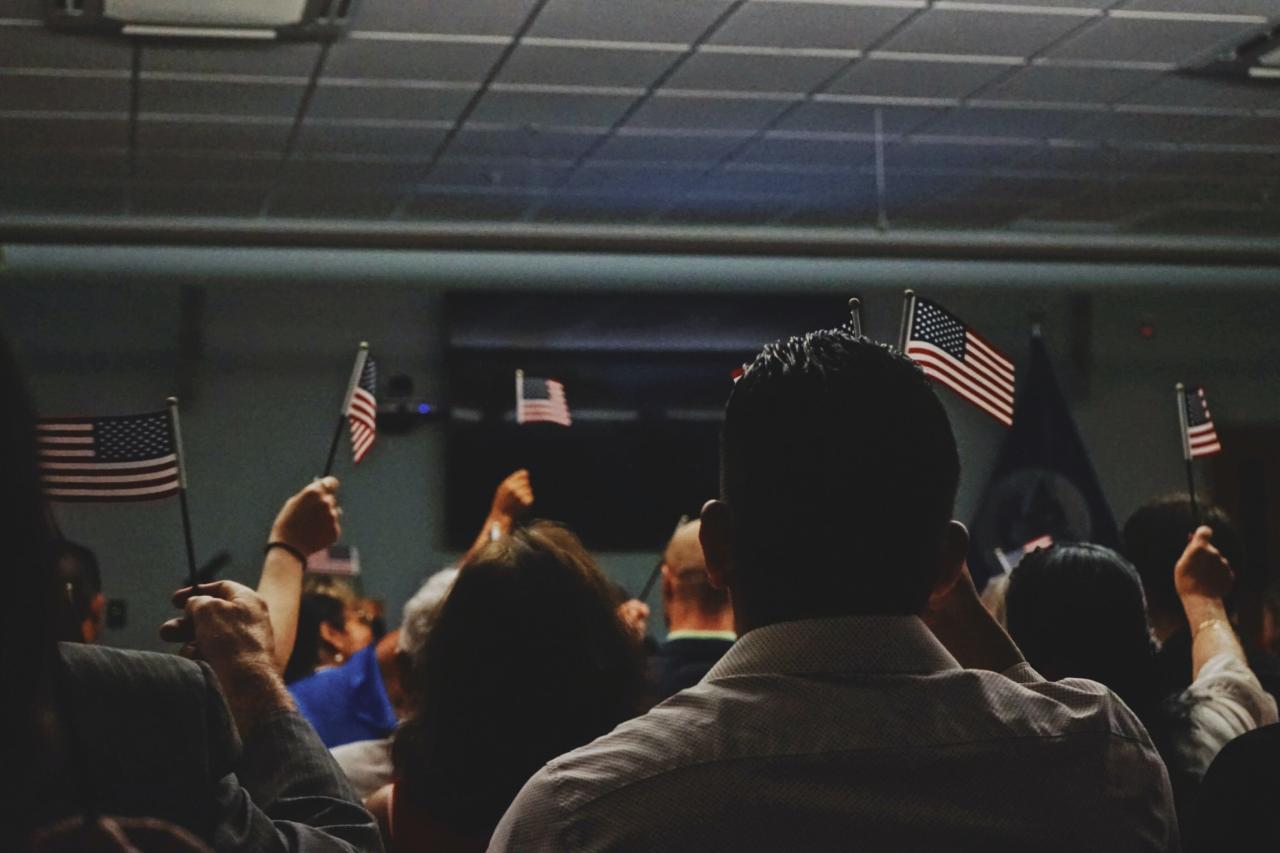 An image of a group of people facing opposite the camera waving small American flags indoors.