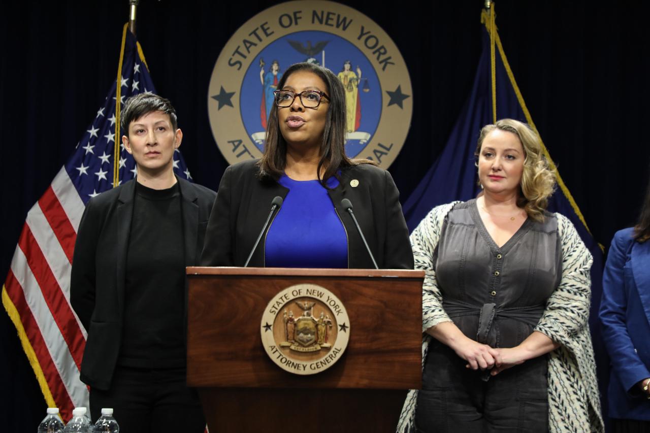 New York Attorney General Letitia James stands at a podium with two individuals standing close behind her. She appears to be giving a speech. 