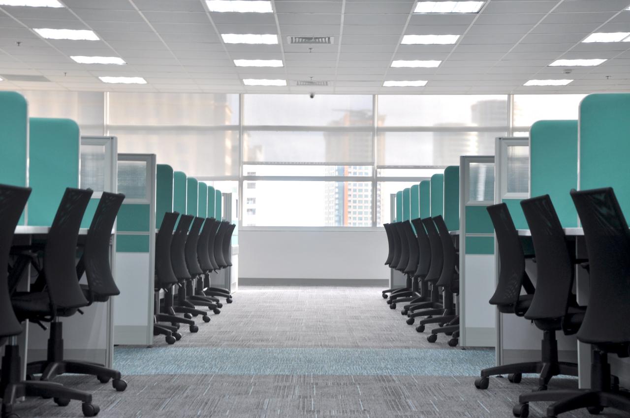 An image with a row of cubicles on both sides. The cubicle walls are aqua colored and the rolling desk chairs are black.
