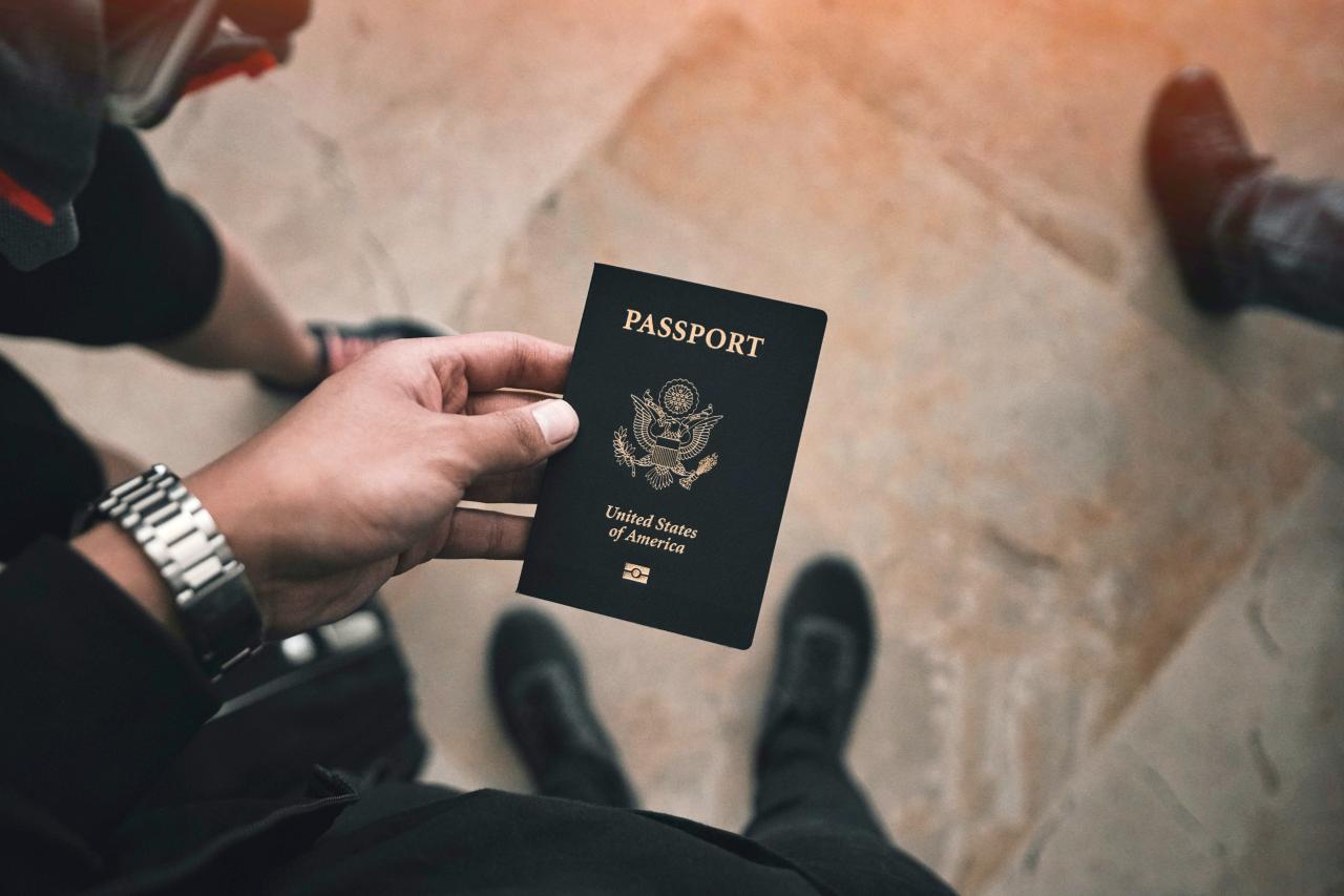 An image of a left hand holding a USA passport. Behind the passport you can see the floor and shoes of the person holding the passport and also of the surrounding people