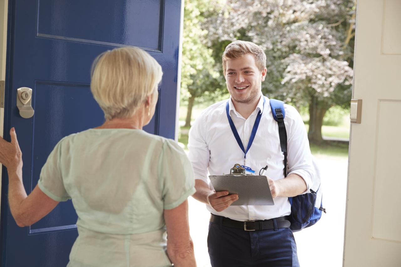 An image of an older woman opening her blue front door to speak with a man in a white button down and blue lanyard holding a clipboard