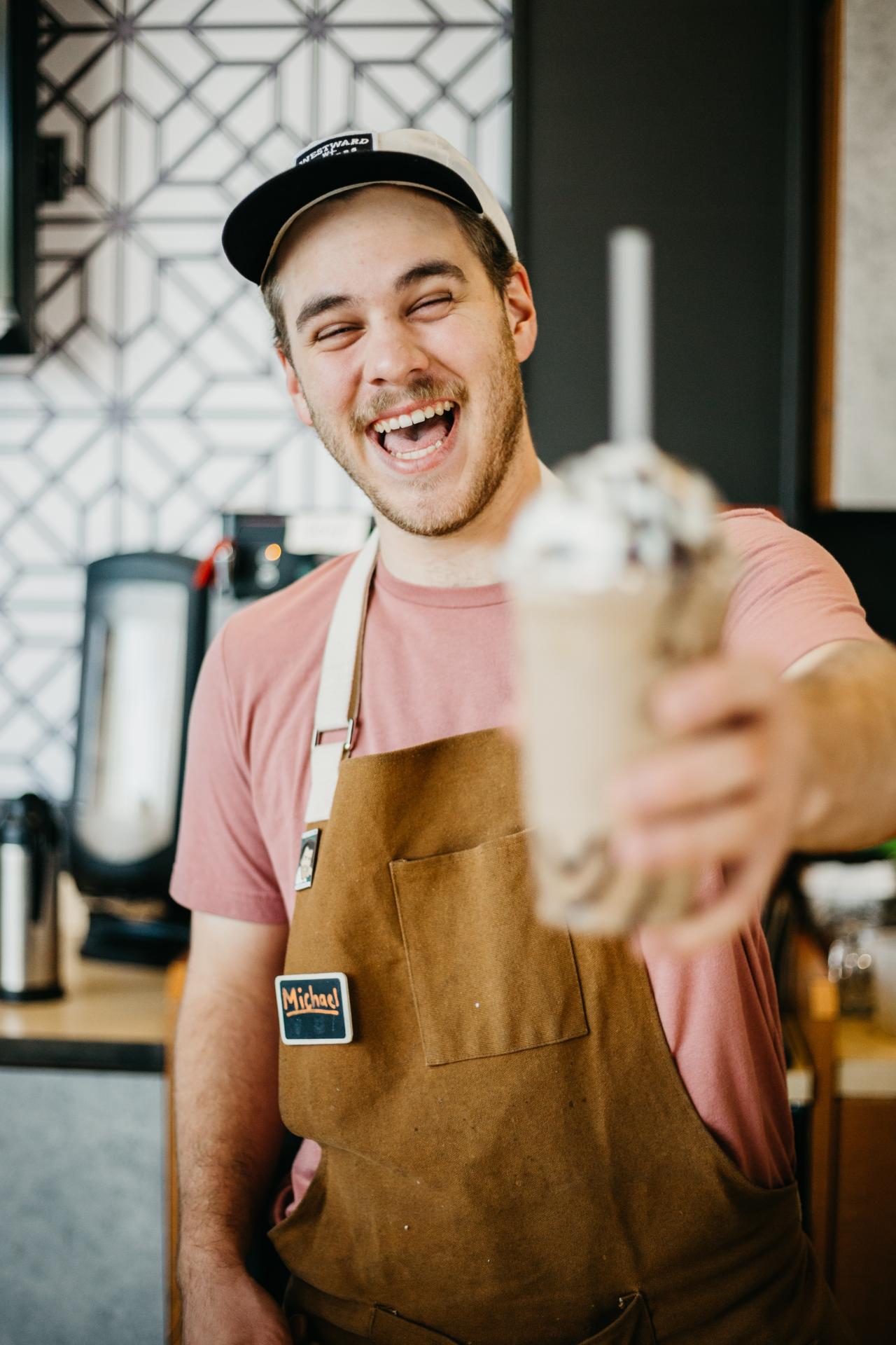 A food working holding a brown drink with whipped cream and chocolate towards the camera. The worker is smiling and wearing a brown apron, pink t-shirt, and a hat.