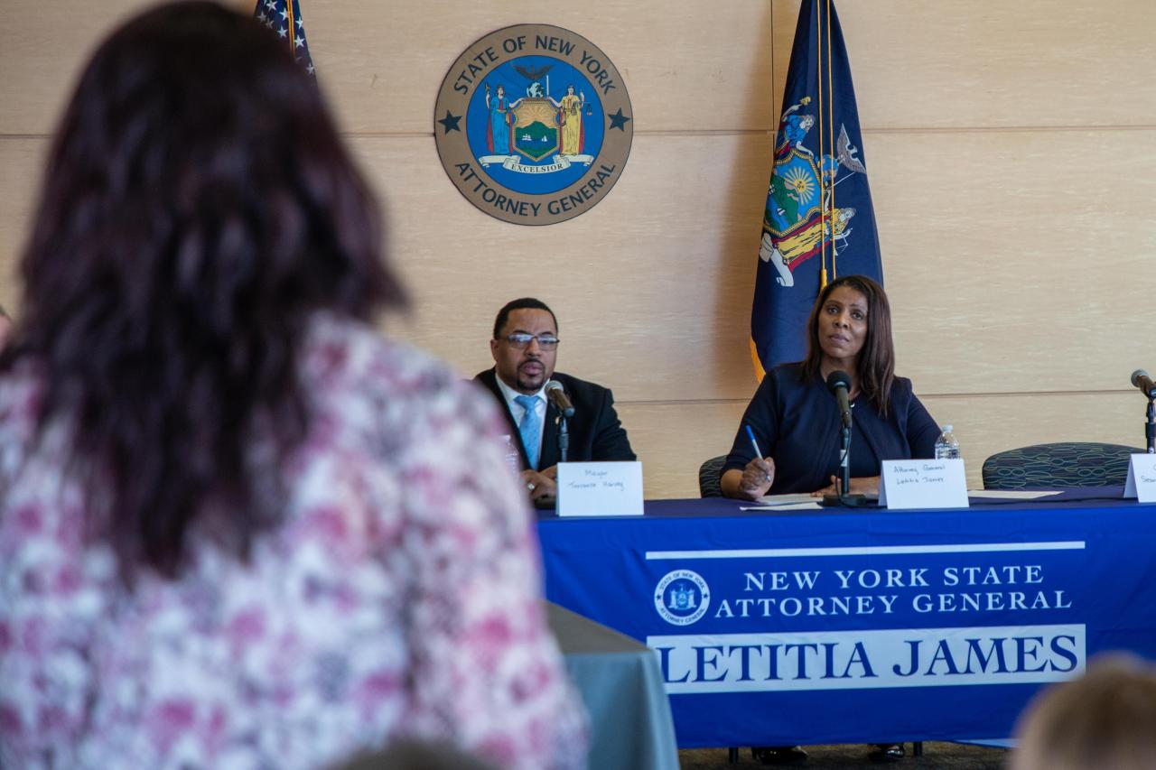 Attorney General James sits behind a table with her name on a banner draped across it. She appears to be taking notes and listening to a member of a seated crowd addressing her, who can be seen out of focus in the left foreground of the image. 