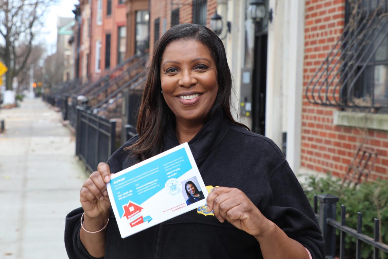 Letitia James stands on the sidewalk in front of a row of brownstones, holding a pamphlet to give to residents. 