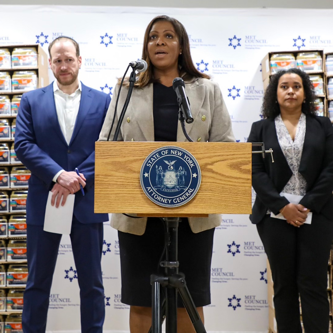 AG James at a podium with two people and a stack of baby formula 
