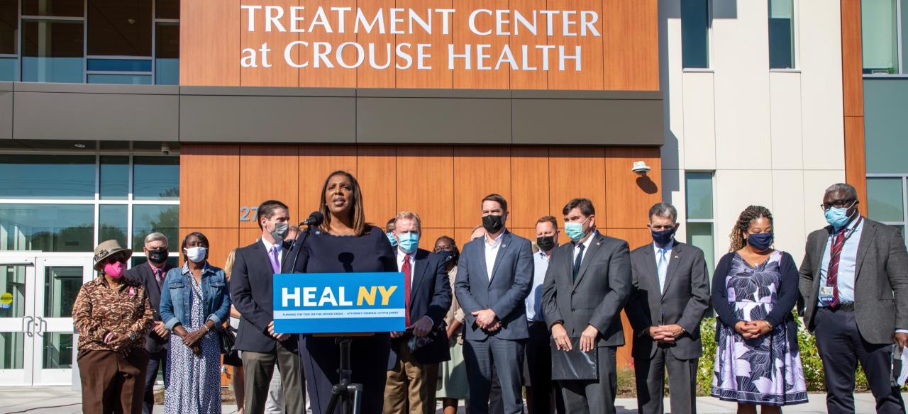 AG James at a podium with a group of people that and sign that reads HEAL NY