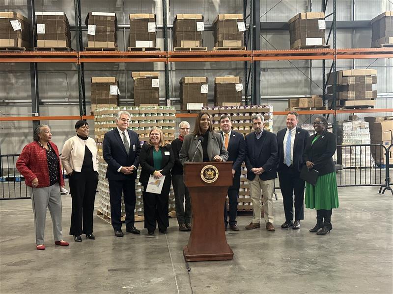 AG James at a podium with a group of people and stacked baby formula boxes behind her